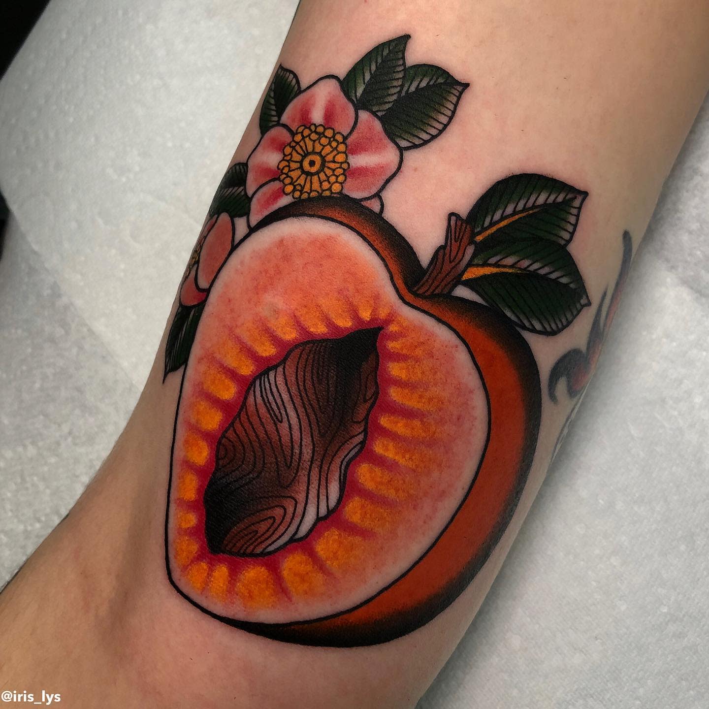 Tattoo of a halved peach with a pit, surrounded by flowers and leaves on an arm